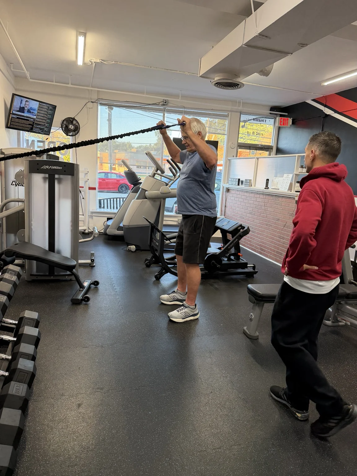 Client raising a resistance band overhead while a trainer watches his form.